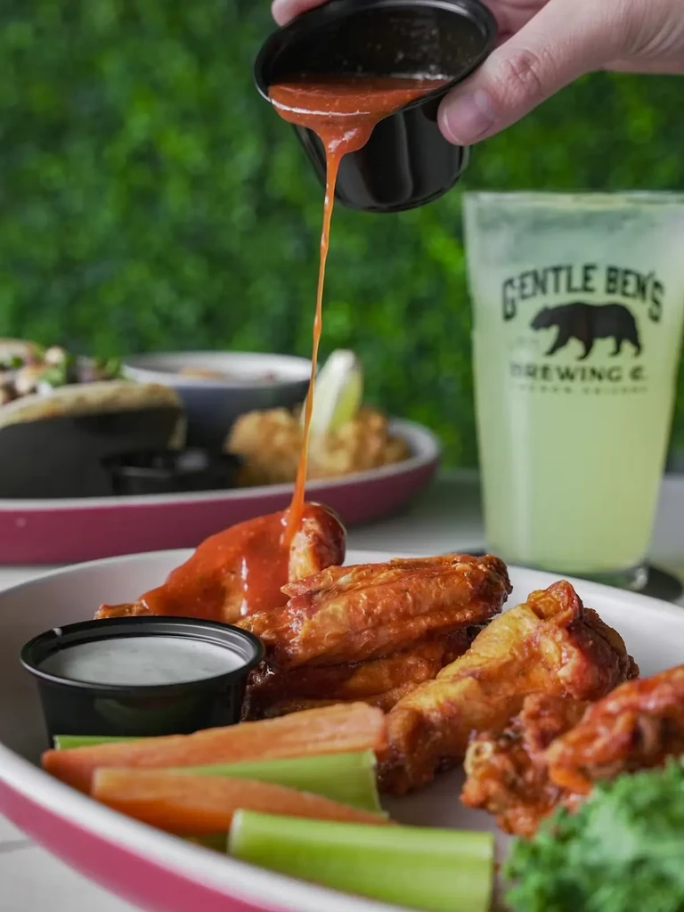 Hand pours hot sauce on crispy chicken wings, with dipping sauce and veggies.