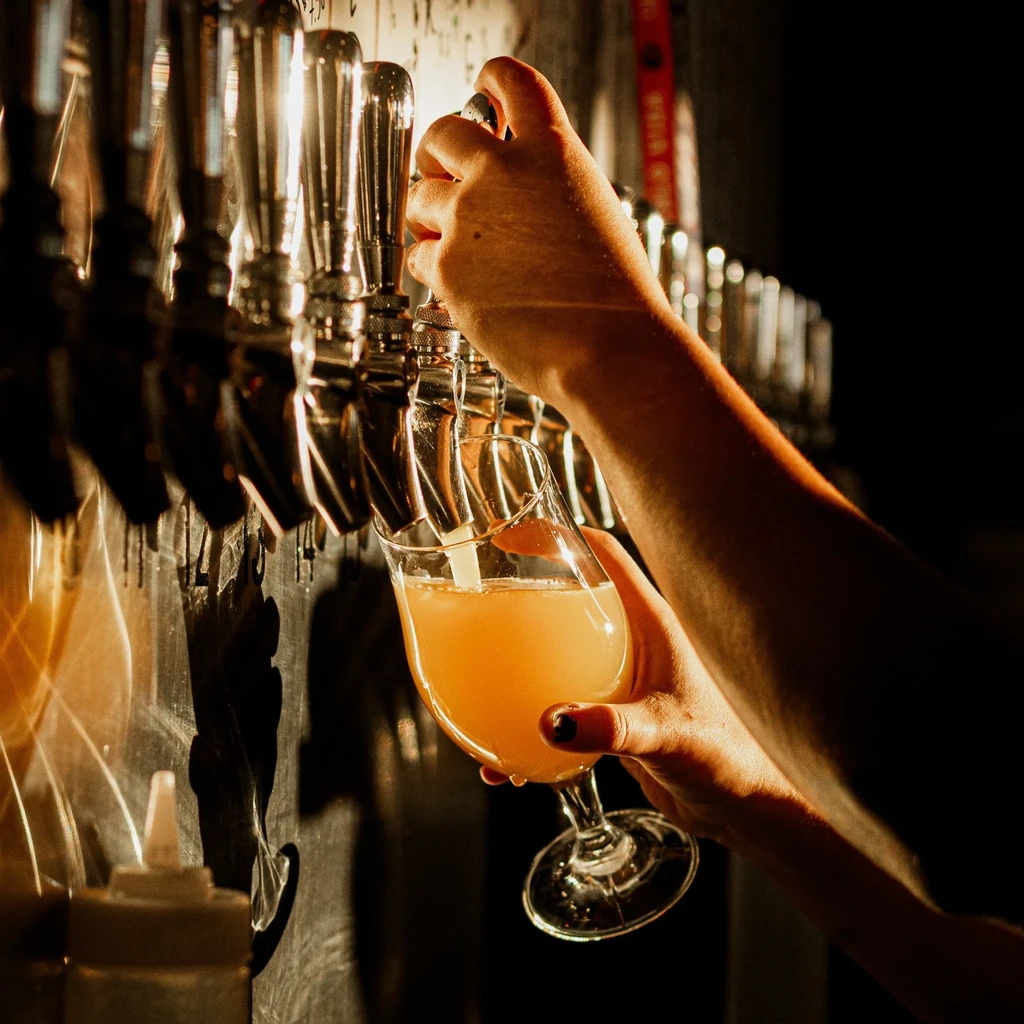 Hands pouring amber beer from a silver tap into a stemmed glass.