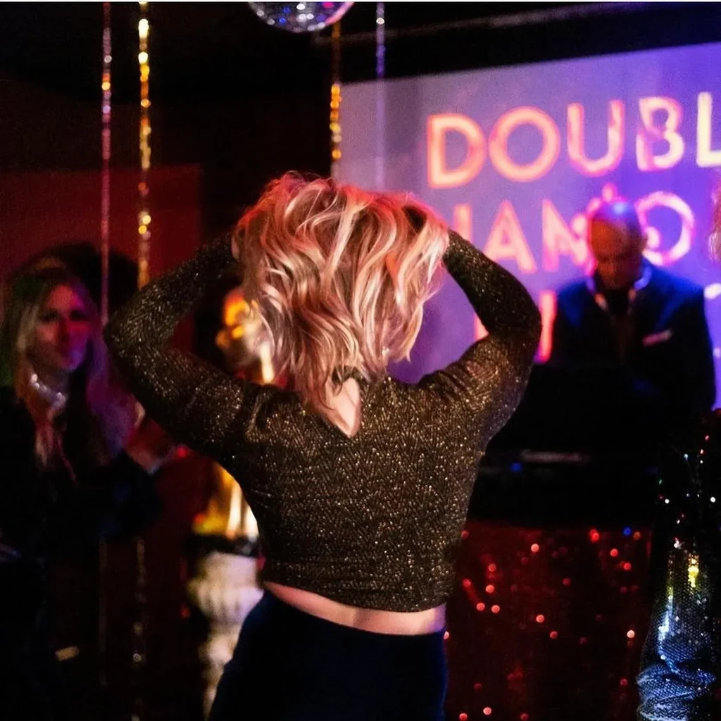 Person with blonde hair and sparkly top dancing in a lively club.