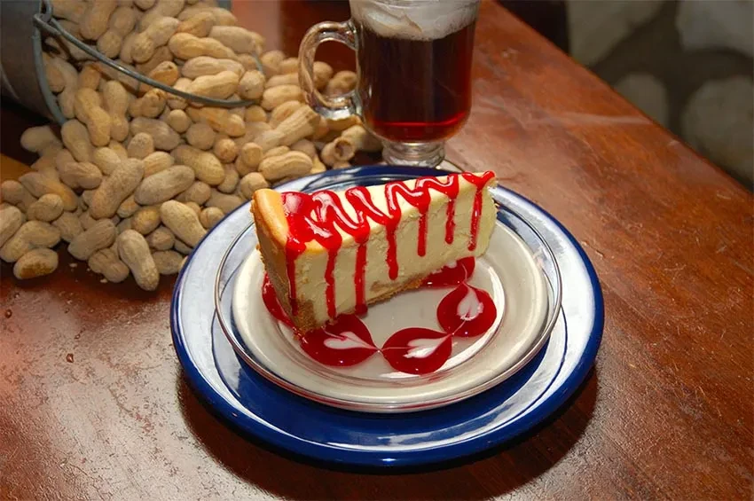 Cheesecake with red sauce, spilled peanuts, and beverage on wooden table.