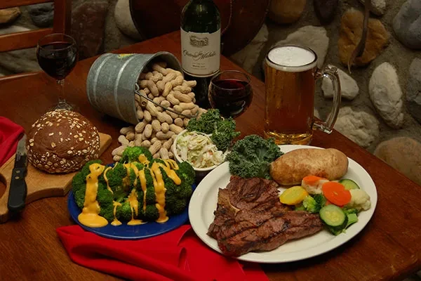 Rustic table of steak, vegetables, bread, wine, beer, and peanuts.