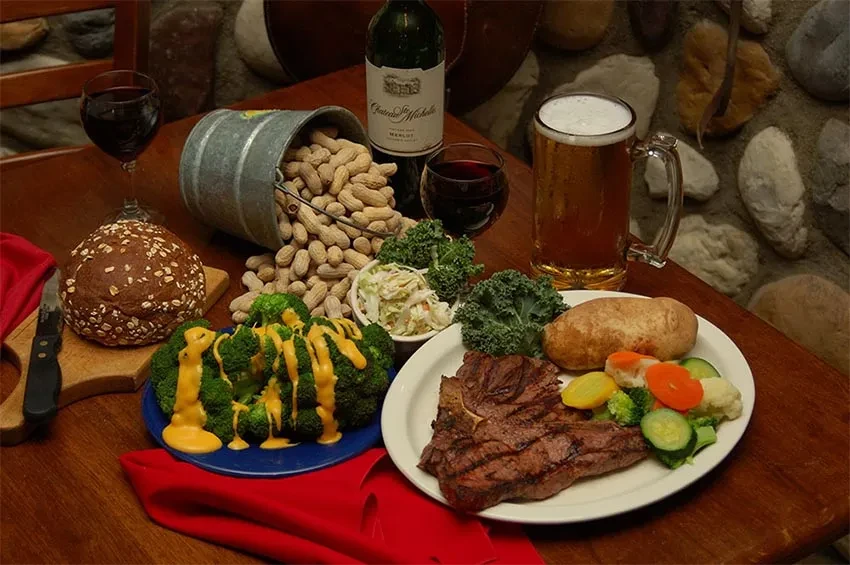Grilled steak, vegetables, bread, wine, beer, and peanuts on a rustic table.