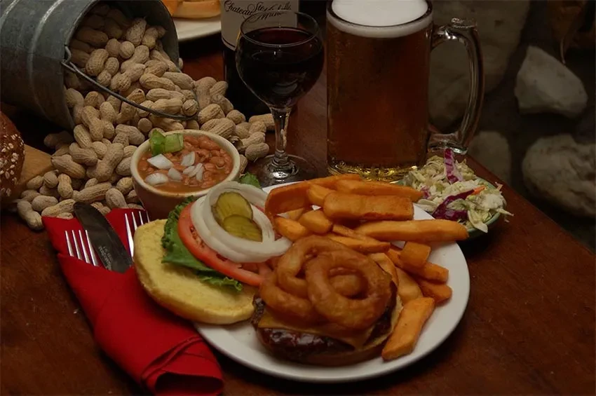 Open cheeseburger, fries, onion rings, beer, wine, peanuts on rustic table.