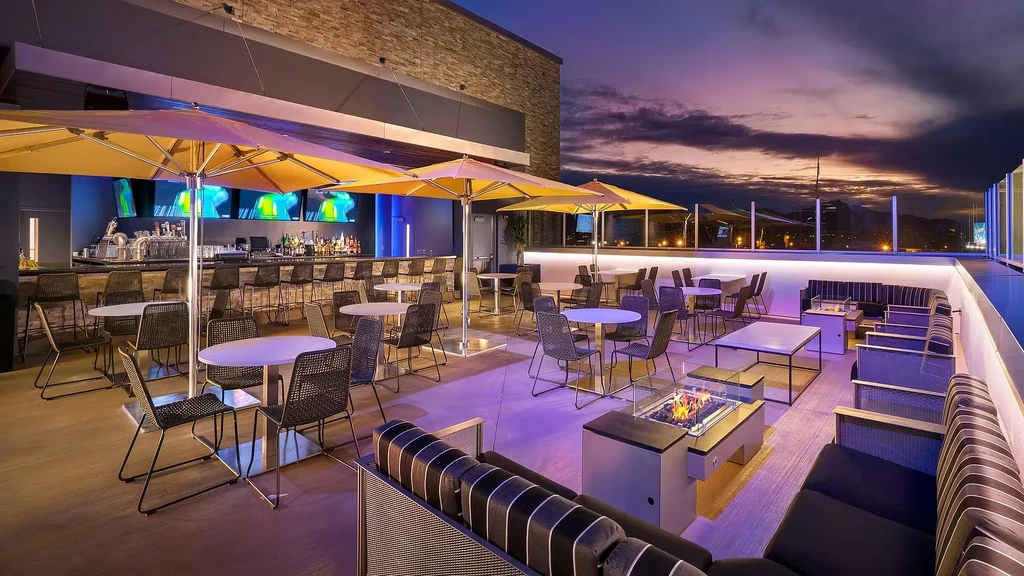 Rooftop bar at dusk with blue-lit bar, fire pits, and city view.