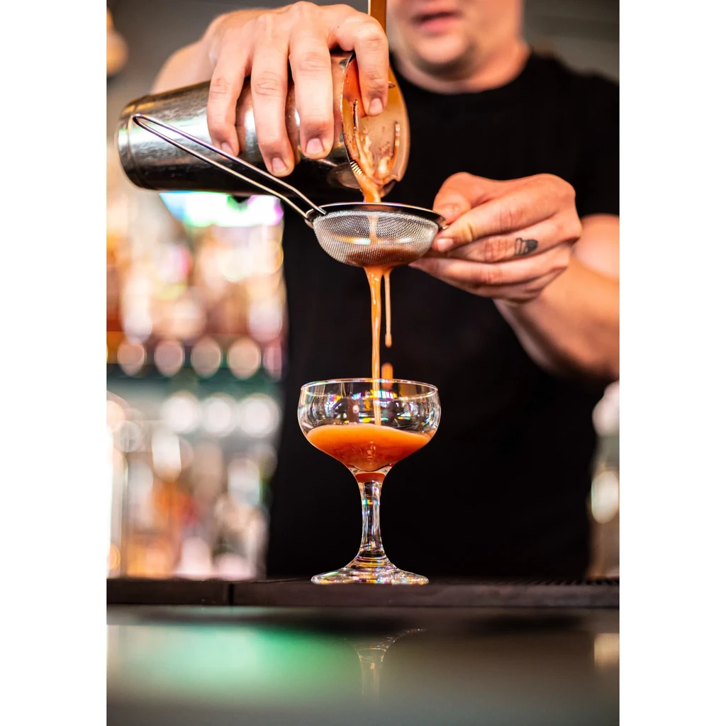 Bartender pouring a reddish-orange cocktail into a coupe glass through a strainer.