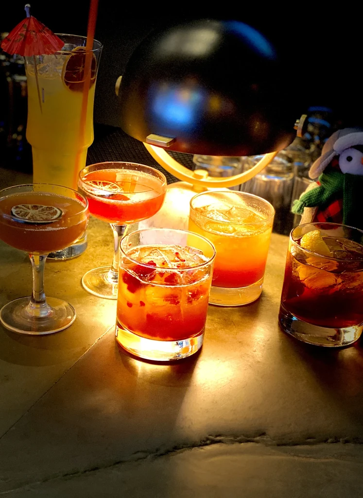 Five colorful cocktails with garnishes on a dimly lit bar.