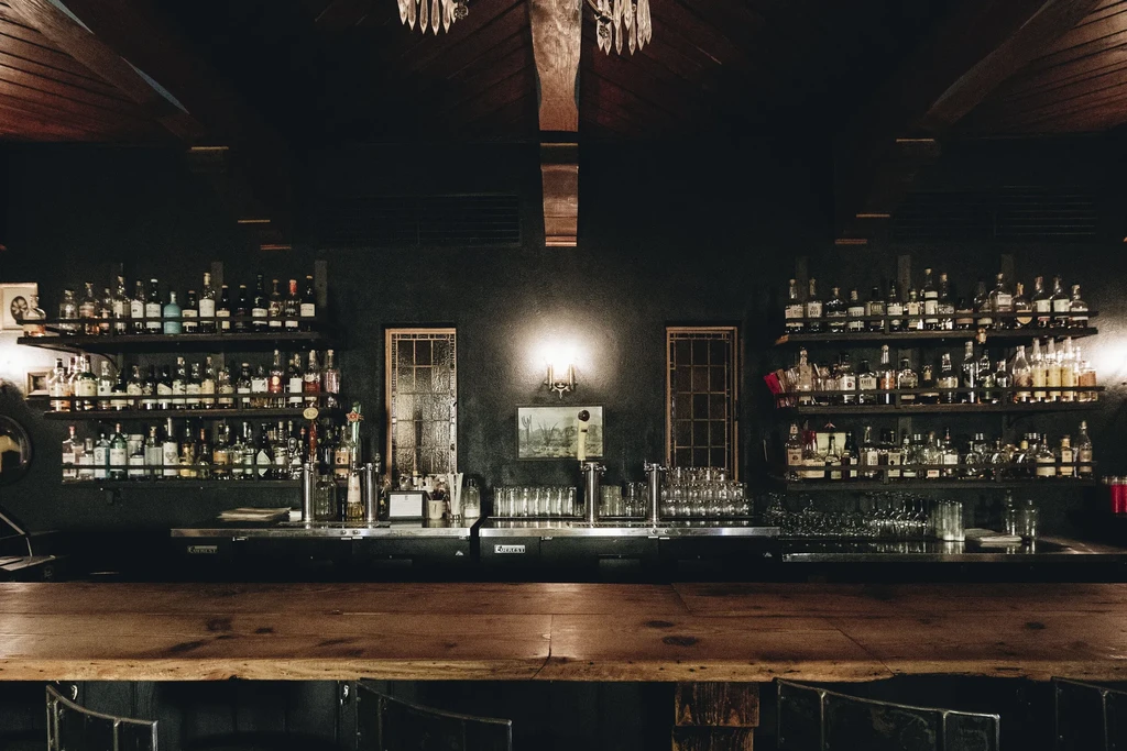 Dimly lit bar interior with rustic counter, stocked shelves, and desert painting.