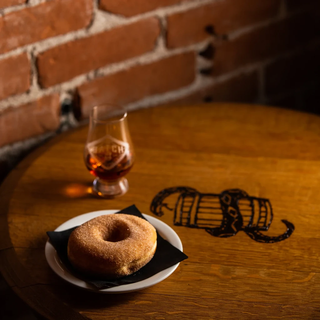 Cinnamon sugar donut, whiskey in 'BATCH' snifter on logo-etched wooden table.