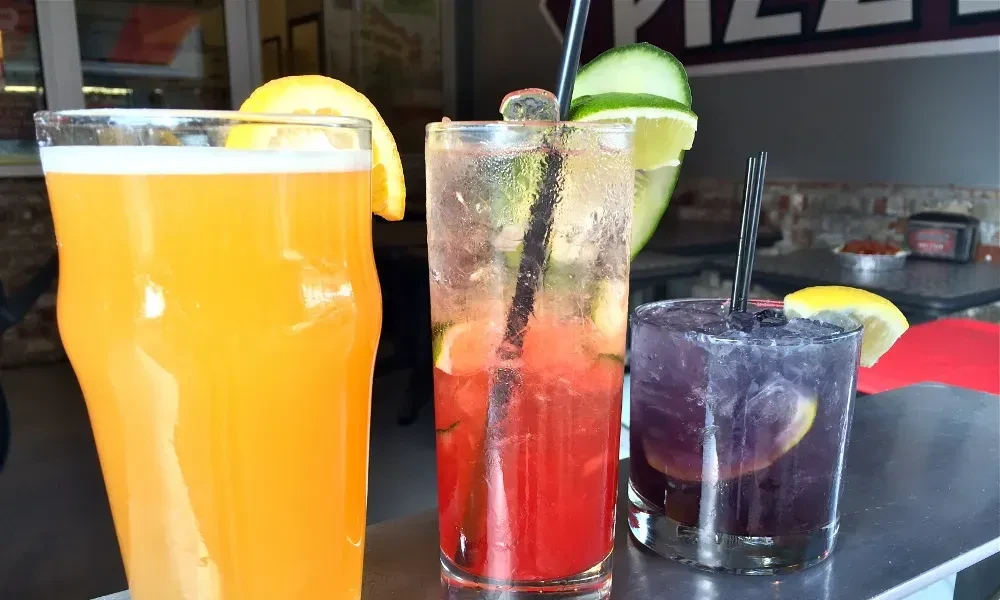 Orange, pink, and purple drinks with ice and garnishes on a bar.
