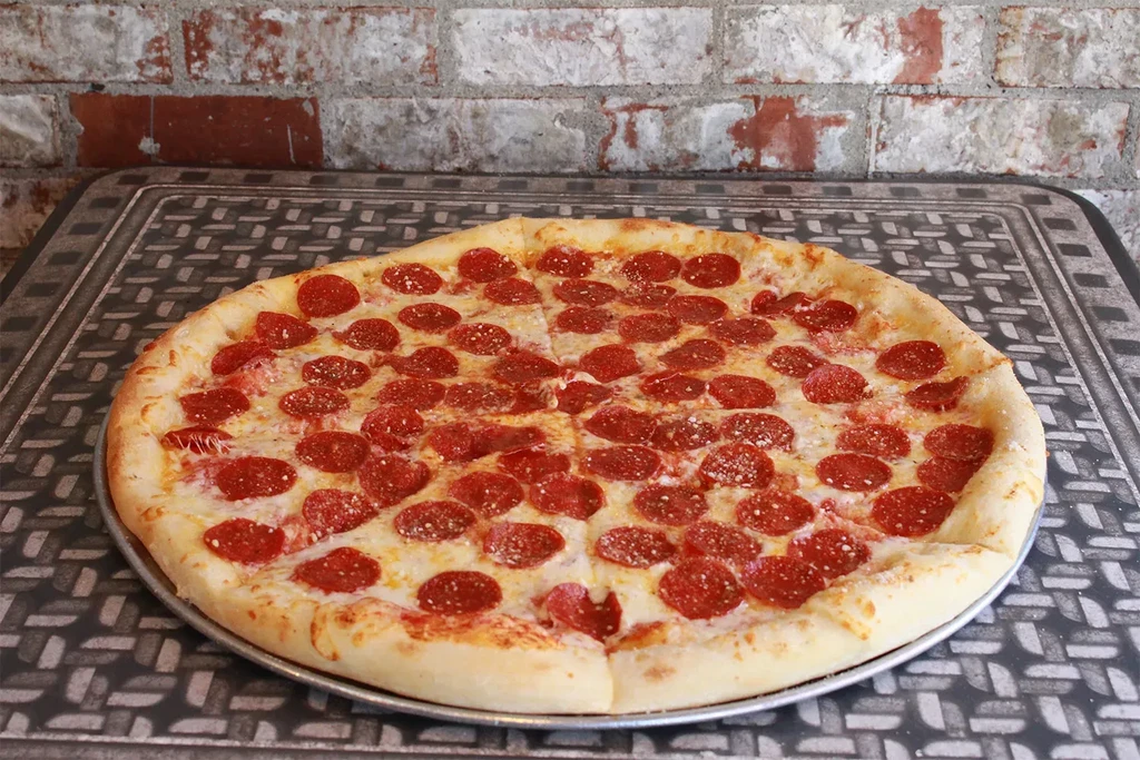 Whole pepperoni pizza on silver pan, dark table, rustic brick wall.