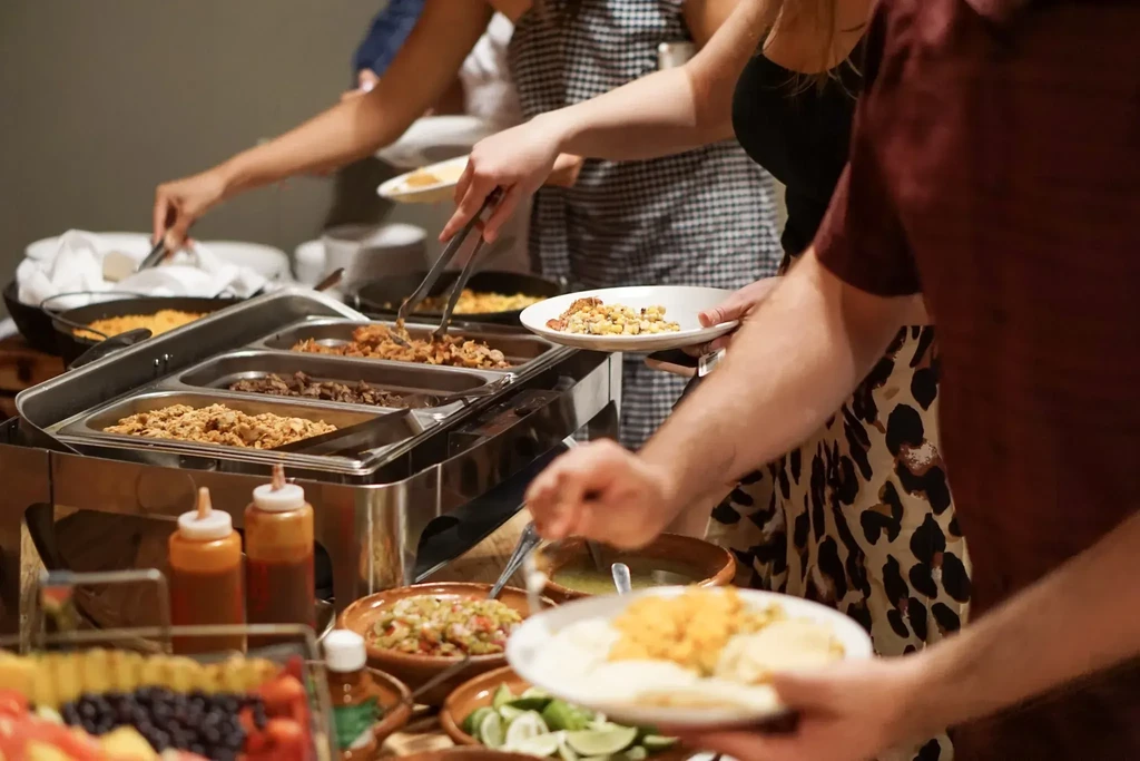 Hands serving diverse dishes from a buffet of rice, tortillas, fruit.
