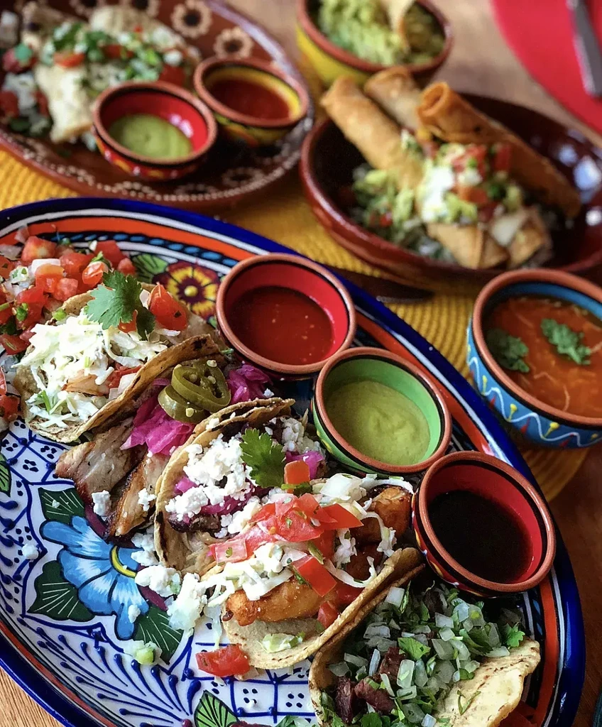 Colorful Mexican food spread featuring tacos, salsas, and flautas on ceramic dishes.