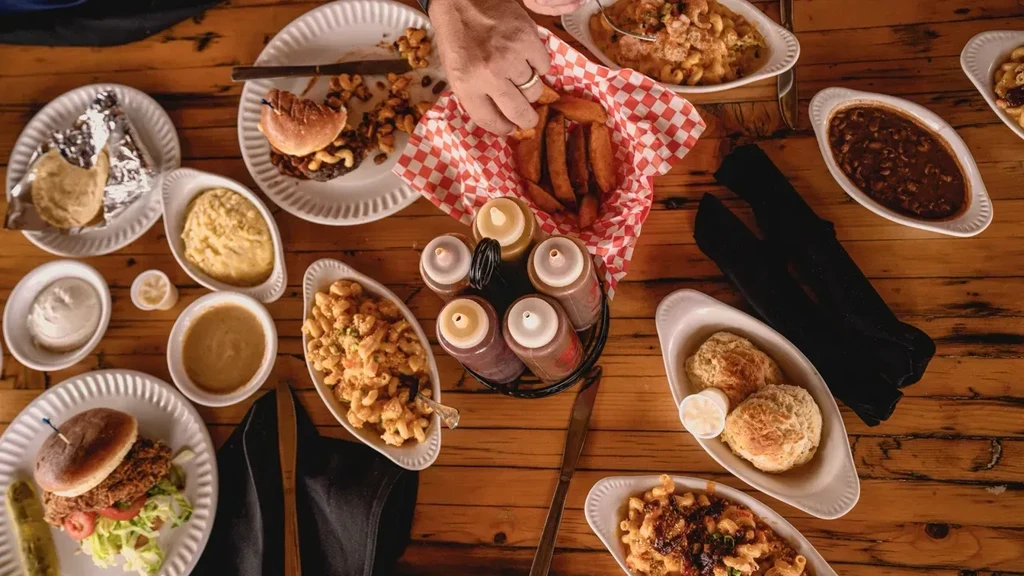 Overhead: Hand reaching for burgers, mac and cheese, fries on wooden table.
