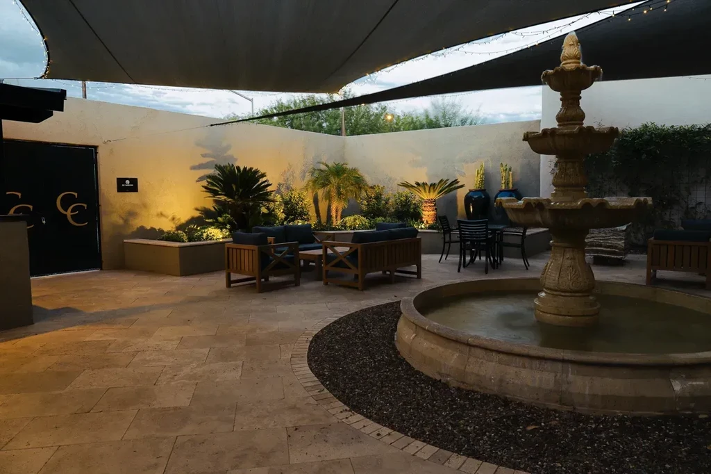 Evening patio with stone fountain, lush plants, outdoor seating, string lights.