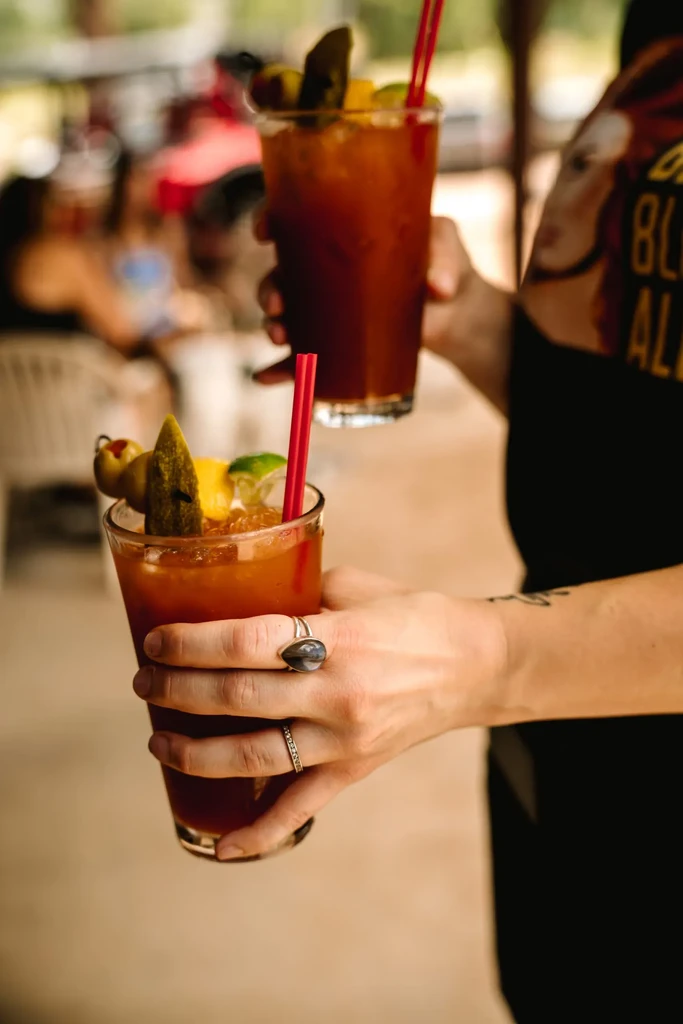 Two hands hold cocktails garnished with olives, pickles, and lime.