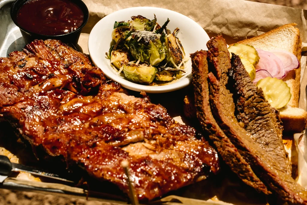 Close-up barbecue platter on tray: ribs, brisket, Brussels sprouts, bread, pickles.