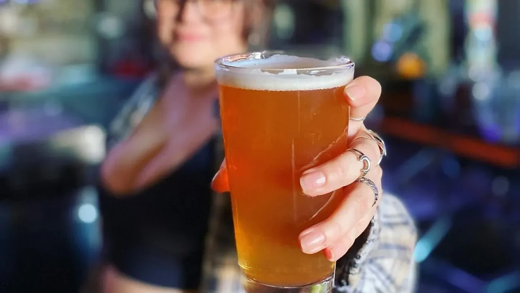 Ringed hand holding a glass of amber beer, blurred bar background.
