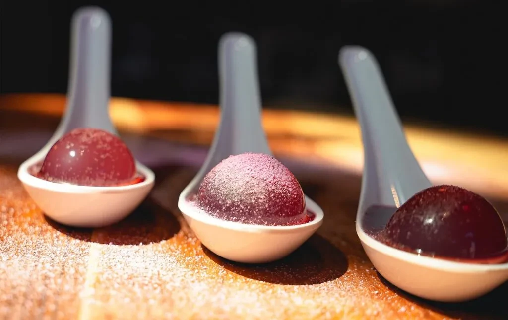 Three spherical red jellies in white spoons on a powdered wooden surface.