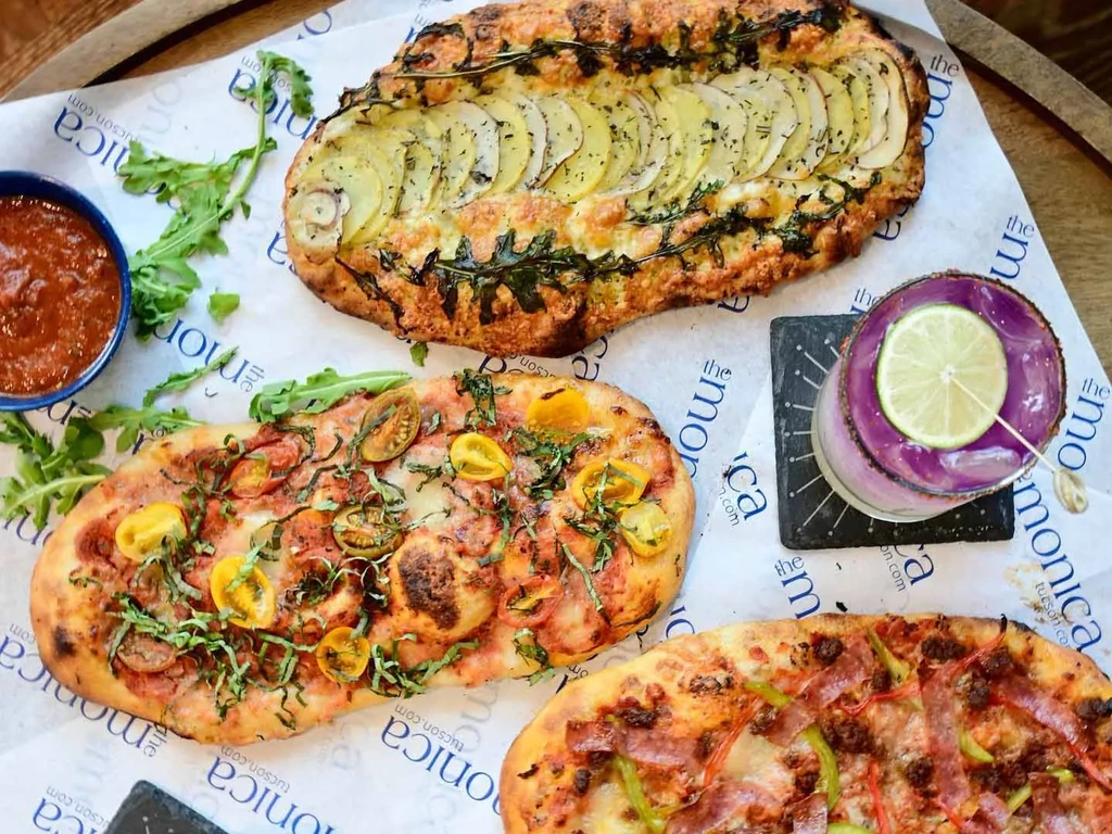 Overhead: three flatbreads, purple cocktail, dip, and arugula on branded paper.