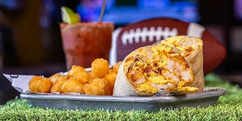 Breakfast burrito, tater tots, Bloody Mary on turf with football.