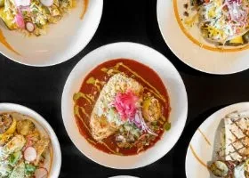 Overhead: Five colorful Latin American dishes on white plates, dark table.