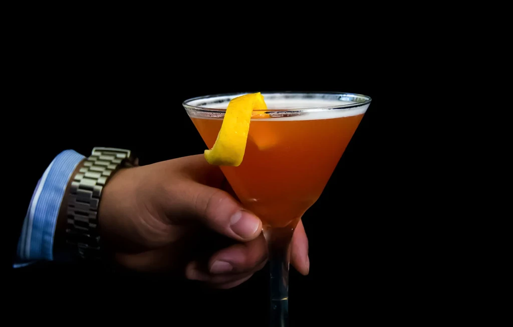 Hand holding amber cocktail with lemon peel in martini glass, black background.