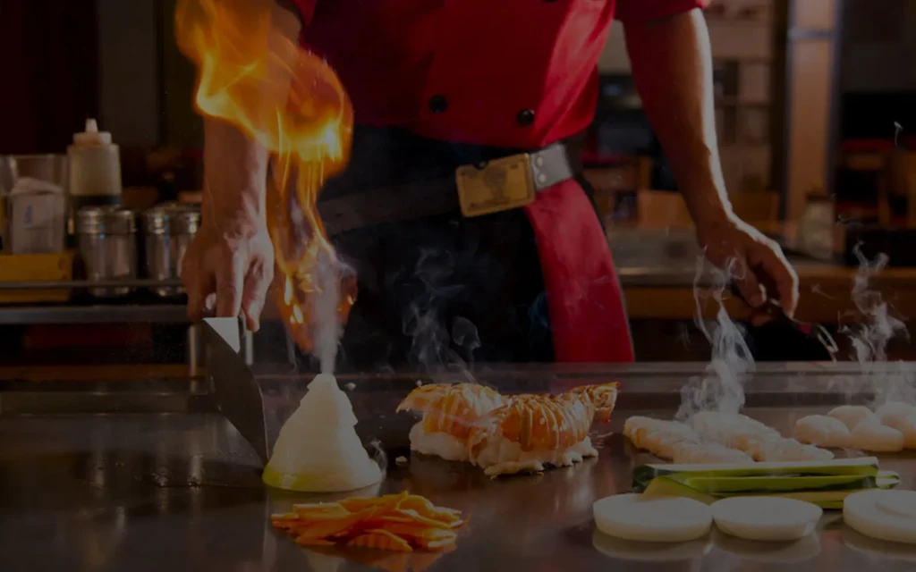 Hibachi chef cooks lobster, vegetables on grill with a prominent onion flame.