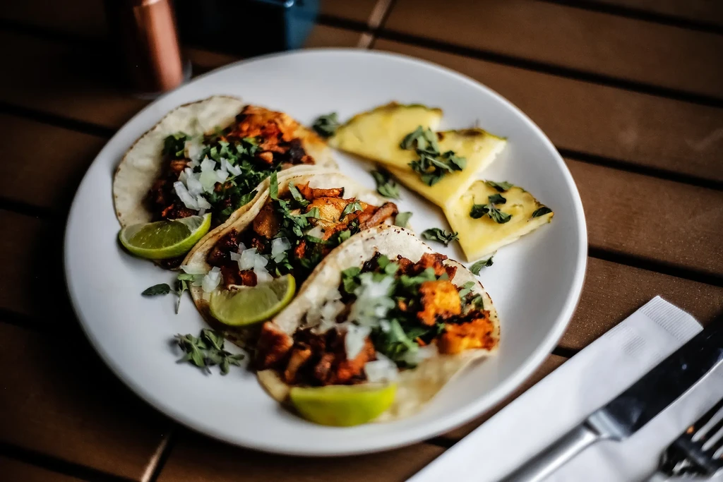 Three tacos with lime, cilantro, and pineapple on a wooden table.