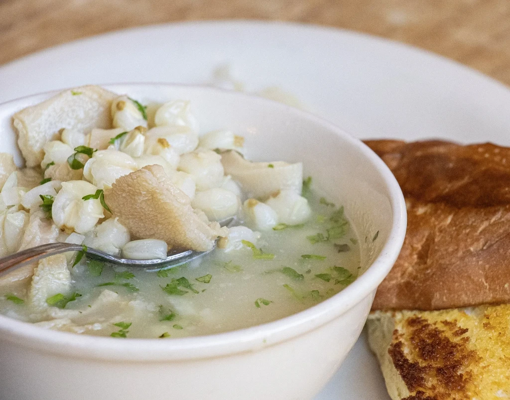 Close-up: bowl of hominy and meat stew with spoon and bread.