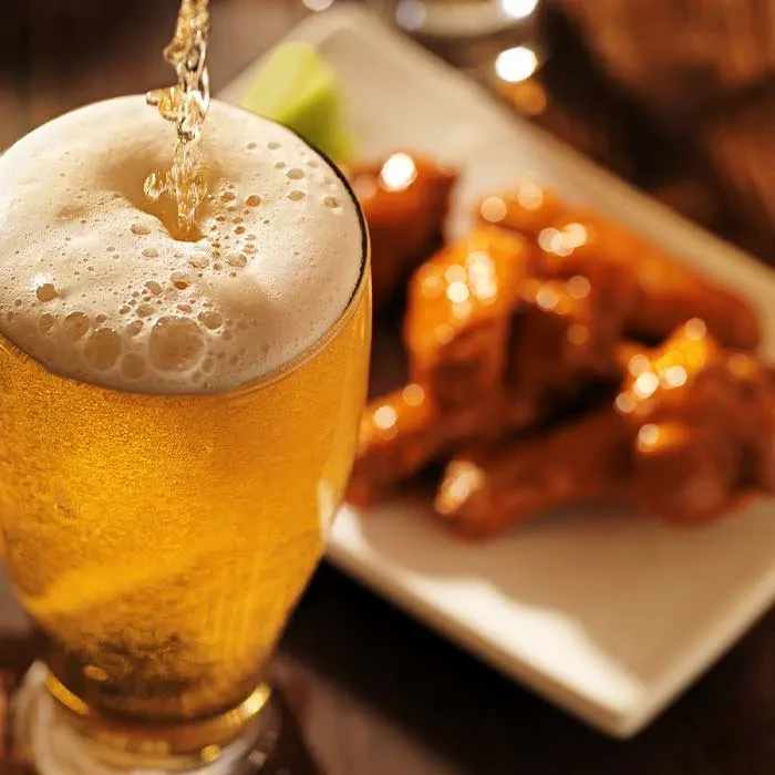 Golden beer actively poured with a frothy head, blurred chicken wings.