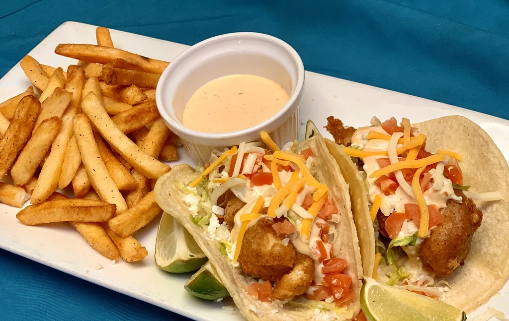 Two crispy fish tacos, golden fries, and dipping sauce on a white plate.