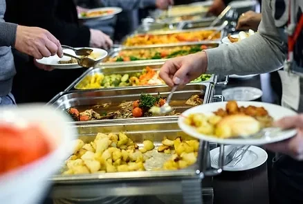 Hands dish roasted potatoes, meat, and vegetables from a vibrant buffet.