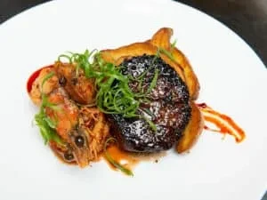 Elegant dish with seared protein, two shrimp, green garnish, and red sauce.