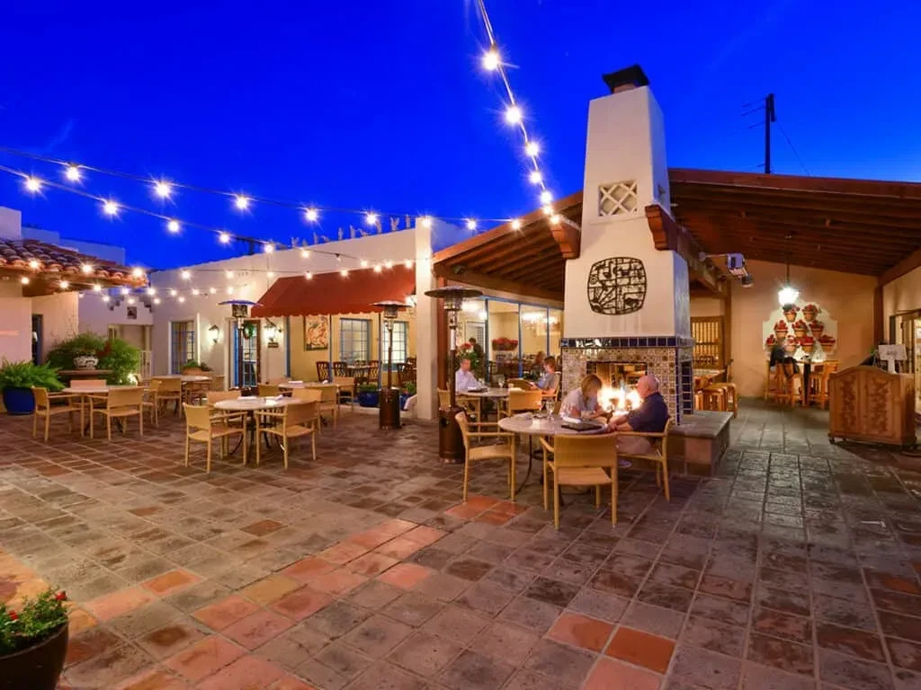 Restaurant patio at dusk with diners, glowing lights, and stucco fireplace.