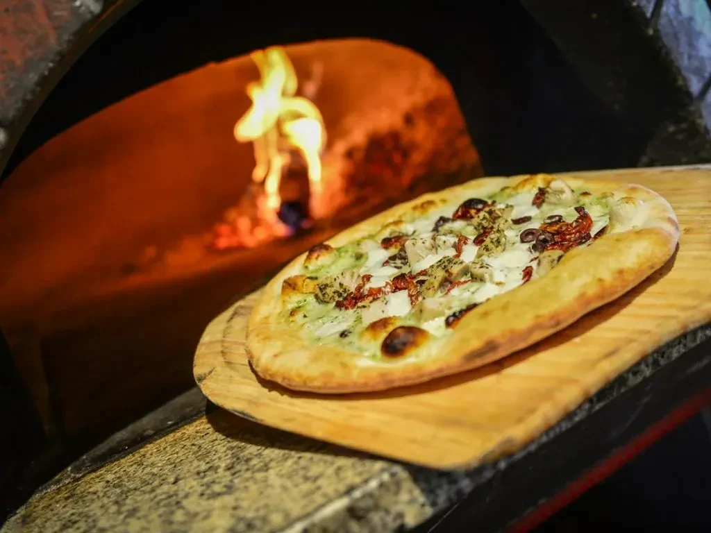 Wood-fired oval pizza with chicken and sun-dried tomatoes on a peel.