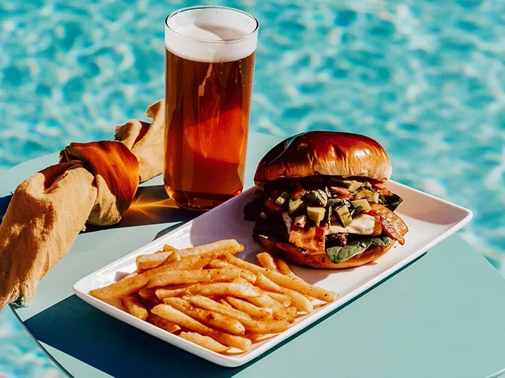 Gourmet burger, fries, and beer served outdoors by turquoise water.