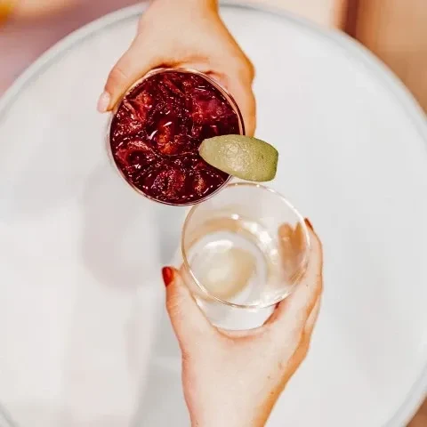 Hands holding one dark red drink and one empty glass tumbler.