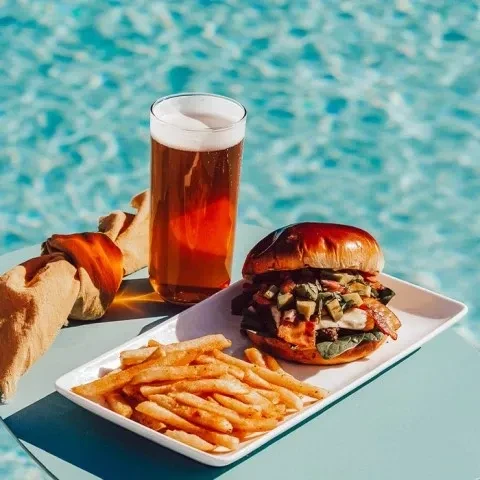 Burger with bacon, avocado, fries, and beer by a pool.