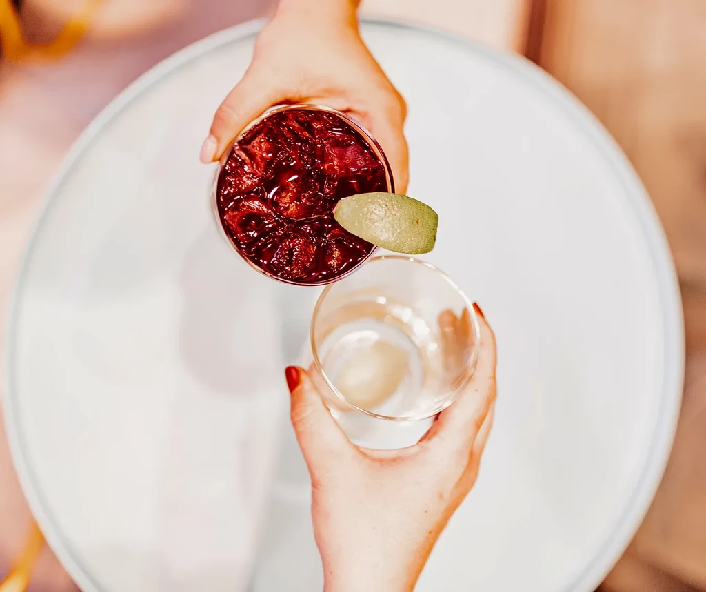 Overhead: Hand holding red cocktail; other hand holds an empty glass.