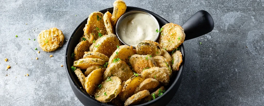 Black bowl of golden fried pickles with herbs and dipping sauce.