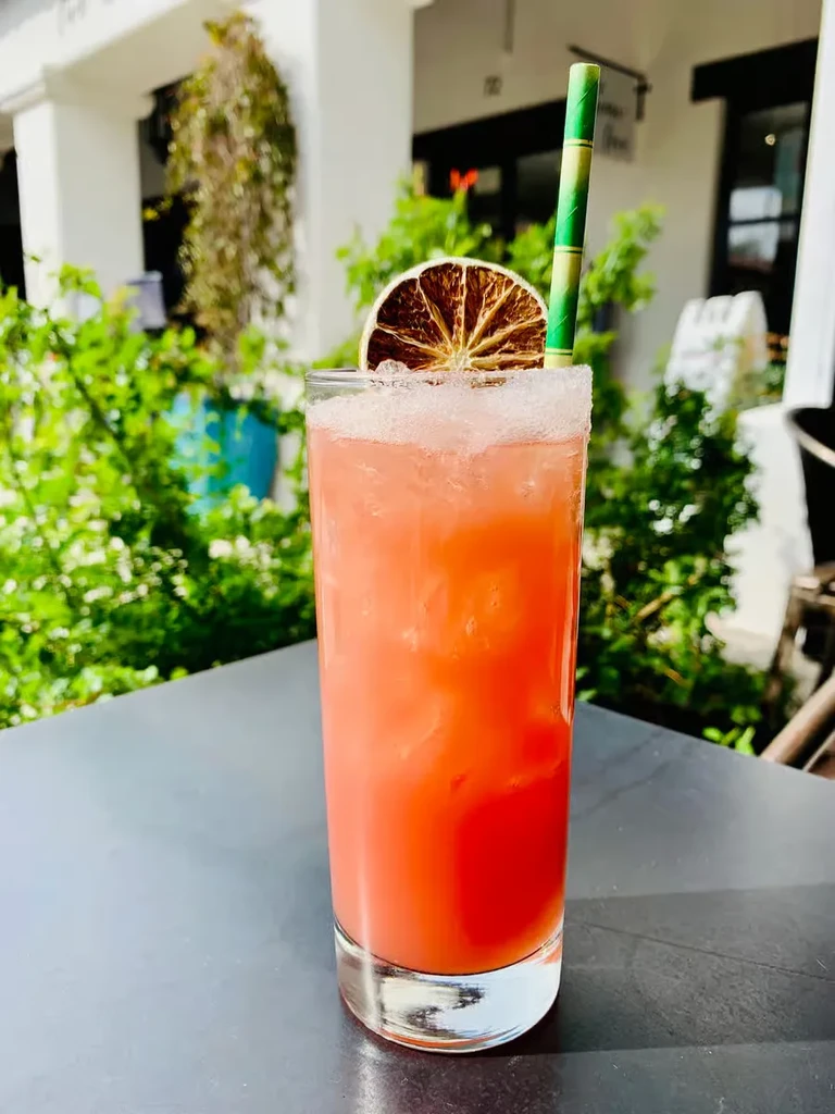Reddish-orange cocktail with citrus slice, straw, and sugar rim outdoors.
