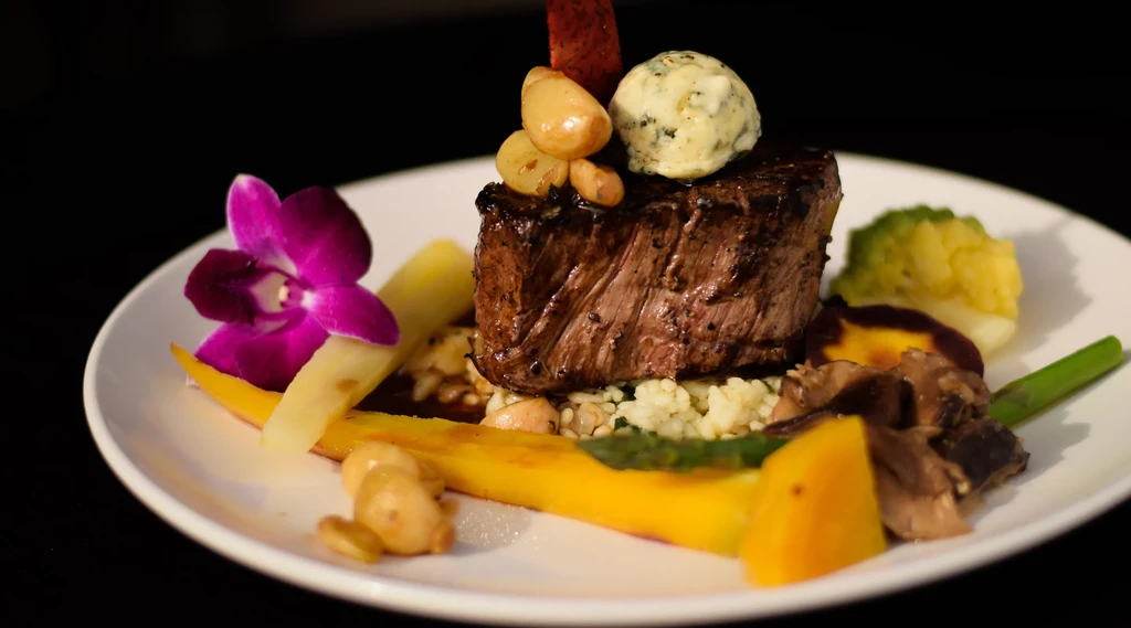 Plated seared meat, roasted garlic, herb butter, vegetables, and an orchid.