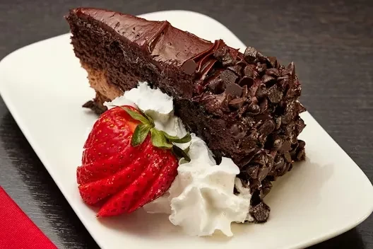 Slice of chocolate cake with ganache, chocolate chips, whipped cream, and strawberry.
