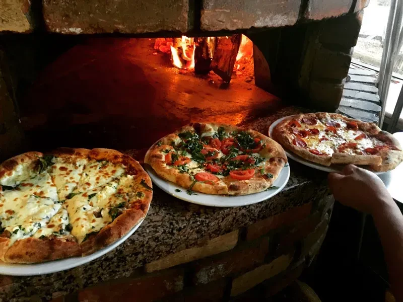 Three assorted pizzas, one held, in front of a glowing brick oven.