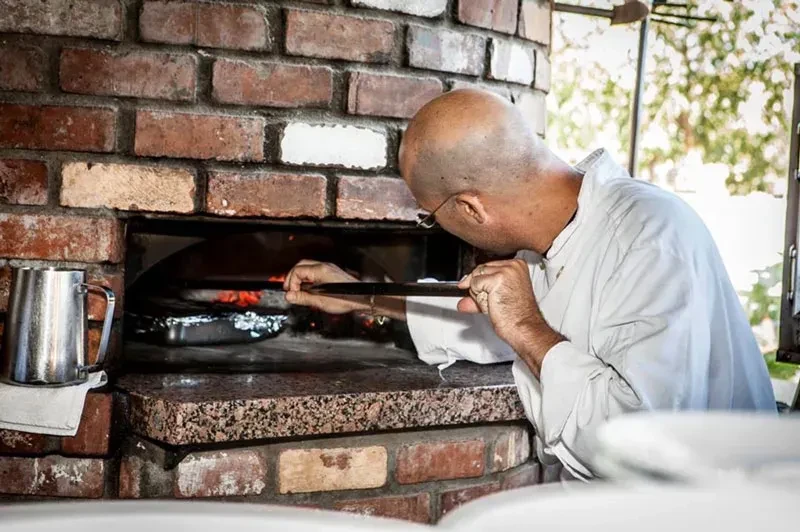 Bald chef with glasses uses tool inside a brick wood-fired oven.