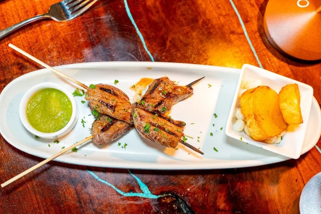 Grilled meat skewers, green sauce, roasted potatoes on rustic wood table.