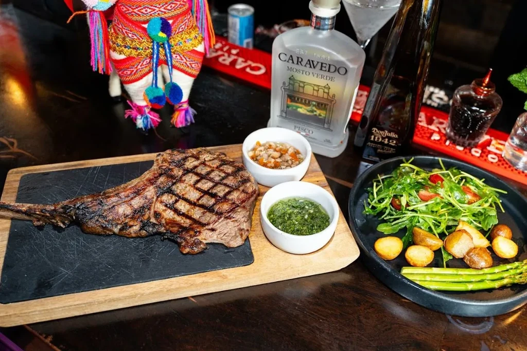 Grilled tomahawk steak, sides, drinks, and llama on a dark bar.