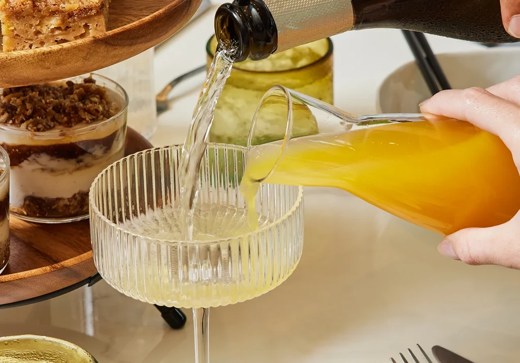 Hand filling mimosa into coupe, desserts on a wooden tray.