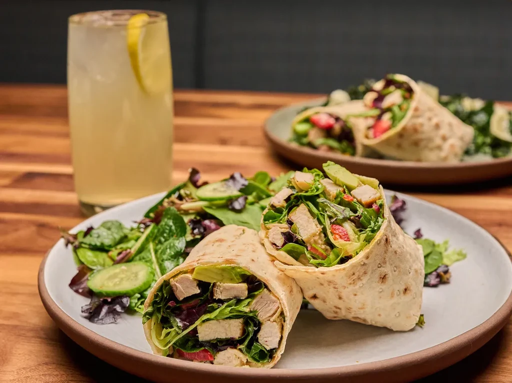 Two grilled chicken wraps, cut in half, with salad and lemonade.
