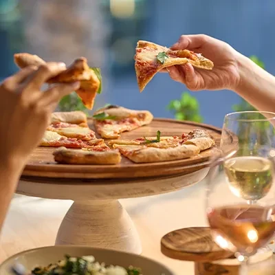 Hands take pizza slices from a wooden stand, with wine.